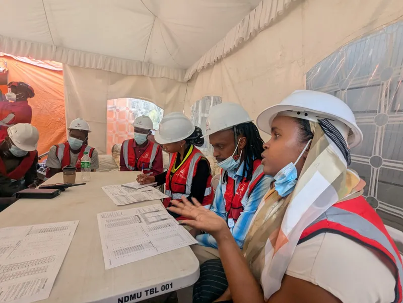 AAK's Mulika Mjengo team in hard hats and red AAK vests at a table of printed sheets in a tent during the site visit