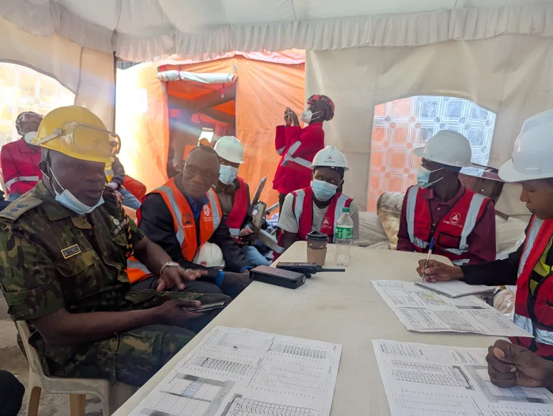 The Mulika Mjengo team at a briefing table in the tent with an officer in camouflage uniform and responders in hard hats