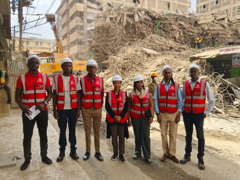 AAK's Mulika Mjengo team in hard hats and red AAK vests in front of the rubble of the collapsed building on Eastleigh 9th Street